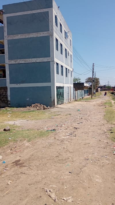 The image depicts an unpaved dirt road in Mosop ward, Kenya, under a clear blue sky. A five-story building occupies the left foreground, painted light blue-gray with lighter concrete horizontal bands delineating floors and vertical bands at corners. Multiple rectangular windows, some ajar, are visible on its facade. The ground floor features a large green corrugated metal gate and an adjacent entry door. A pile of bricks lies on the patch of dry grass at the building's base. The dirt road, littered with assorted debris including plastic and paper, extends into the distance, flanked by sparse dry vegetation. On the right, a utility pole supports multiple overhead power lines. A white and black goat stands on the road's right shoulder in the mid-ground. Further back, a small group of individuals is present near low-rise structures. In the far distance, two figures, one distinct in reddish attire, walk away from the viewer along the road. Distant trees and power line towers are visible against the horizon.