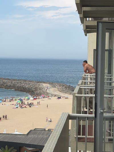 A man is leaning over a balcony railing, looking out at a beach scene. The image is taken from a high vantage point, looking down at the beach and the ocean. The man is shirtless and wearing swim trunks. He is holding something to his mouth, possibly a cigarette or a vape.
The beach is crowded with people, many of whom are sitting on towels or under umbrellas. There are also several boats anchored in the water. The ocean is calm and blue. The weather appears to be sunny and warm. The location is Espinho, Portugal. The image seems to be taken in the late afternoon or early evening, as the sun is starting to set.
The overall mood of the image is relaxed and peaceful. The man on the balcony appears to be enjoying the view, and the people on the beach are enjoying the sun and the sea. The image is a snapshot of a perfect summer day at the beach.