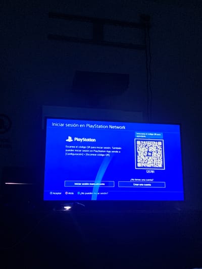 The image shows a television screen displaying a PlayStation login screen. The screen is blue and features the PlayStation logo and text in Spanish. The text on the screen indicates that the user needs to scan a QR code to log in to the PlayStation Network. A QR code is displayed on the screen, along with a code "125781". Below the QR code, there are options to "Iniciar sesión manualmente" (Log in manually) and "¿No tienes una cuenta? Crear una cuenta" (Don't have an account? Create an account). At the bottom of the screen, there are controller button prompts: "X Aceptar" (X Accept), "O Atrás" (O Back), and "△ ¿No puedes iniciar sesión?" (△ Can't log in?). The television is in a dark room, with some ambient light casting shadows on the wall behind it. The overall scene suggests a user attempting to log into their PlayStation account. There is no discernible information in the image to place it in El Valle del Espíritu Santo, Venezuela.