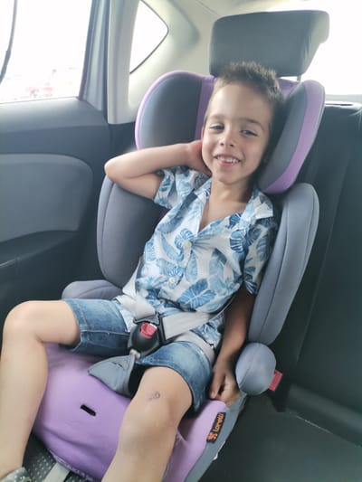 A young boy with short, dark hair and a wide smile is seated in a purple and gray car seat. He is wearing a short-sleeved, blue and white floral print shirt and denim shorts. His left arm is resting behind his head, and his right hand is on his lap. The car seat is positioned in the back seat of a car, with the dark gray car interior visible on the right side and the window and outside view on the left. The overall lighting suggests it is daytime. A small, orange and black "Lorelli" logo is visible on the side of the car seat. The image was taken in Dobrich, Bulgaria.
