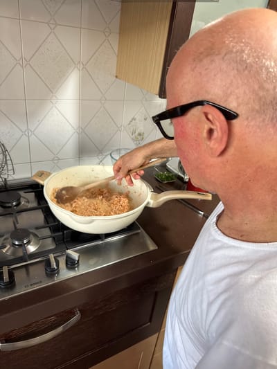A person is cooking in a kitchen. The person is stirring food in a white pot on a gas stovetop with a wooden spoon. The person is wearing a white tank top and glasses. The kitchen has white tile floors and dark brown cabinets. The stovetop is branded "Bosch." The location is a kitchen in Aprilia, Italy.