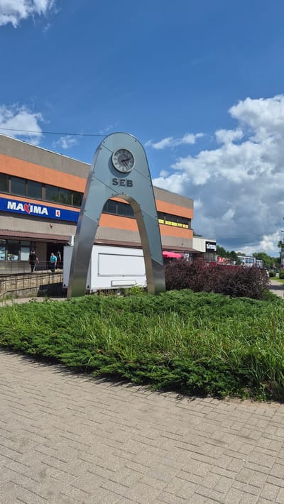 The image captures a sunny day in Daugavpils, Latvia, with a clear blue sky dotted with white, puffy clouds. The scene is a street view featuring a modern building with orange and gray facades. Prominently displayed on the building is a large, gray, arch-like structure housing a clock. The clock face shows the time is approximately 10:50. The text "SEB" is visible next to the clock, indicating a business. Below the arch, the blue and white text "MAXIMA" is emblazoned on the building, identifying a store.

A well-maintained green hedge runs along the side of a paved walkway, which occupies the right side of the frame. The lighting suggests it is daytime, likely late morning or early afternoon. The overall atmosphere appears to be calm and ordinary, a typical day in a commercial area. Two individuals are partially visible near the entrance of the Maxima store, suggesting people are going about their daily activities. The weather appears pleasant and warm, judging by the bright sunshine and clear skies. The architectural style of the building and the visible signage provide strong cues about the location being a developed urban area in Latvia.