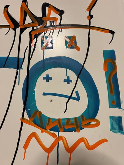 The image appears to be a close-up of an abstract artwork or graffiti on a white surface. The artwork consists of various shapes and lines in the colors blue, orange, and black. A blue circle with a white center containing a number "1" and a plus sign is a prominent feature. Other elements include orange question marks and squiggle lines. Black lines are randomly dispersed across the image. There is no information provided in the prompt to suggest the location of this image is Bečići, Montenegro.