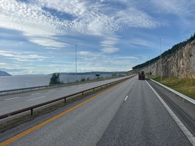 The image shows a view from a vehicle traveling on a highway in Espa, Norway. The highway runs alongside a body of water on the left and a rocky, tree-covered cliff face on the right. The sky is blue with scattered white clouds. A truck is visible in the distance on the right lane of the highway. The road has multiple lanes and is marked with white and yellow lines. Guardrails line the sides of the road. The environment appears to be a scenic, coastal area.