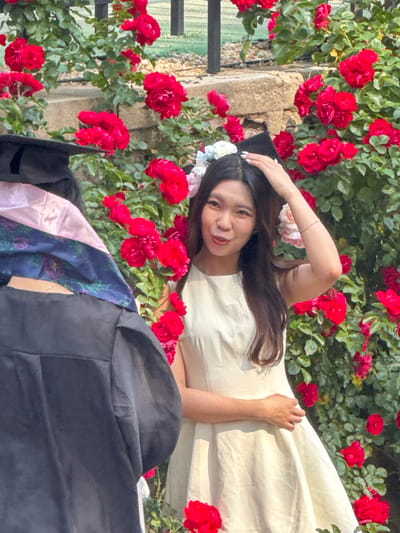 This media file captures a joyful graduation portrait of a young woman taken outdoors in Zhongtun, China, during bright daylight hours, likely midday or afternoon.

The primary subject is a young East Asian woman, positioned lying on her back amidst a dense bed of vibrant red roses and green foliage. She is looking up at the camera with a wide, happy smile and relaxed expression. Her long, dark hair is spread around her head, and she wears a light-colored (possibly cream or white) dress. A black graduation cap, adorned with small white and light blue floral embellishments, rests on her head. Her left arm is bent, with her hand gently touching the cap, while her right arm rests on her dress.

The scene is dominated by numerous clusters of bright red roses, creating a lush, celebratory backdrop. Visible behind the roses and the woman are parts of a textured stone wall. In the lower foreground, a black academic gown and a patterned academic stole, featuring blue fabric with floral designs and hints of lighter colors like pink or lavender, are draped, indicating the graduation event. The bright lighting suggests a sunny day. There is no visible text in the image.