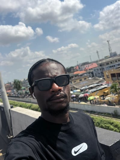 The image is a vertical, selfie-style photograph, rotated 90 degrees clockwise, featuring a man outdoors in an urban environment. The subject is a dark-skinned man, captured from the chest up, looking towards the camera. He wears black, rectangular-framed sunglasses that cover his eyes and part of his nose. A short beard or stubble is visible on his chin, and a white earbud is inserted into his right ear.
He is dressed in a plain black crew-neck t-shirt. On the left side of his chest, a prominent white logo is visible, resembling an elongated oval containing a black Nike swoosh.
The background depicts a vibrant urban landscape, identified as Ikeja, Nigeria. Numerous buildings of varying sizes and architectural styles are densely packed, with some showing weathered facades. A body of water or a canal, partially lined with green vegetation, can be seen winding through the cityscape. The man appears to be positioned on an elevated structure, such as a bridge or overpass, providing a vantage point over the city below. The sky is bright blue with scattered white cumulus clouds, indicating a clear and sunny daytime. The overall impression is a casual portrait taken on a bright day in a bustling city.