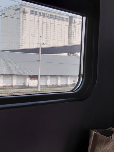 An outdoor shot captures a view from inside a train window, looking out at industrial buildings and railway tracks. In the foreground, the dark interior of the train is visible, with the edge of a brown woven bag in the bottom right corner. Beyond the window, a long, grey, corrugated-roofed building stretches horizontally, its white, plastered walls showing signs of age and weathering. A single, closed wooden door or window is visible on this building. Behind this structure, a much larger, tall, white building with numerous windows runs along the top and is partially obscured by overhead power lines and a dark, elevated walkway connecting to other structures. The area between the buildings is characterized by railway tracks and patches of green vegetation. The sky is overcast and pale. The scene is situated in Prokudskoye, Russia.