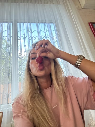 A young blonde woman is taking a selfie with a strawberry held in front of her face, her lips pursed as if to kiss it. She is wearing a pink t-shirt and a silver watch on her left wrist. She is indoors, in front of a window covered by sheer white curtains. The window has a decorative wrought-iron grate and a view of trees and the sky. The background suggests a domestic setting, possibly a home or a cafe. There are no visible landmarks or details that would pinpoint the location to Armavir, Russia, or any specific city.