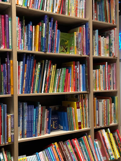 The image displays a close-up view of several bookshelves filled with books. The shelves are made of light-colored wood and are organized into vertical sections. The books are of various sizes, colors, and designs, with many featuring colorful covers. Some books are standing upright, while others are leaning against each other. The lighting in the image is neutral, creating a well-lit but not overly bright environment. The overall scene suggests a bookstore or a library setting. The image does not contain any people or explicit actions. The context of Kommunarka, Russia is not visually represented within the image itself.