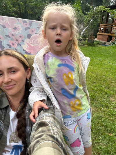 In Udelnaya, Russia, a young girl with blonde hair is captured in a candid outdoor shot, her mouth open in surprise. She wears a tie-dye t-shirt with yellow and blue butterfly-like graphics and grey leggings adorned with colorful hearts, rainbows, and the word "LOVE." To her left, a woman with brown hair braided and wearing a plaid shirt and a cross earring is partially visible, smiling at the camera. They are seated on a bench covered by a floral fabric. The background features a lush green lawn, trees, and a wooden gazebo, suggesting a backyard setting in a rural or suburban area.