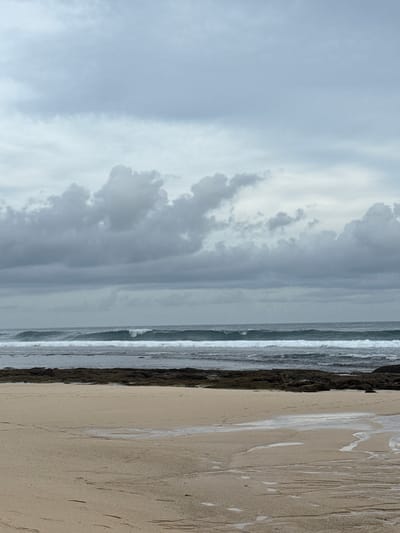 The image depicts a beach scene in Pecatu, Indonesia, under a cloudy, overcast sky. A stretch of wet sand dominates the foreground, marked with patterns likely left by receding waves. To the left of the sand is a dark, rocky area followed by the ocean, with white-capped waves. The sky is filled with heavy gray clouds.