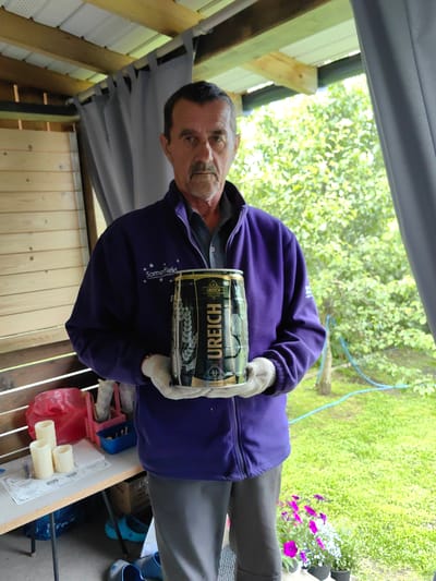 A man wearing a purple jacket and white gloves stands in an outdoor covered area, holding a large can of Ureich beer. The can is dark with gold lettering and features an image of wheat stalks. The man has a mustache and appears to be in his 50s or 60s. To the left of the man, on a table, are three pillar candles of varying heights, a red plastic bag, and a pink plastic organizer with utensils. In the background, there are trees and green foliage, indicating an outdoor setting. The ground is covered in grass, and there are some potted flowers in the lower right corner. The structure he is standing in appears to be a gazebo or a similar outdoor shelter with wooden siding and a corrugated metal roof. The man is facing the camera and holding the can with both hands.