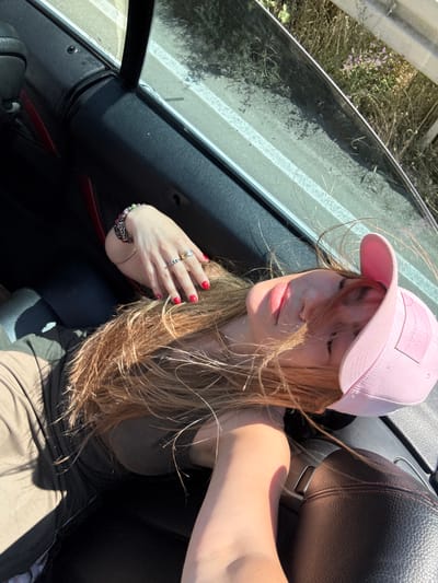 In this image, a young woman with long, blonde hair is pictured sitting in the passenger seat of a convertible car. She is wearing a pink baseball cap and a khaki tank top. Her eyes are closed, and her head is tilted slightly back, suggesting relaxation or enjoyment of the moment. Her hair is blowing in the wind, a common occurrence when the top is down. She is wearing several rings on her fingers and a bracelet on her wrist.
The car's interior is visible, with black leather seats and red stitching on the dashboard. Through the open window, a roadside scene is visible, featuring dry grass, small purple flowers, and a road with white markings, likely indicating a highway or a rural road. The sunlight suggests it is daytime, and the presence of wind implies it might be a breezy day. The context of Dzegvi, Georgia, places the scene in a rural or semi-rural area. The woman's expression and the open-top car suggest a leisurely drive, perhaps a road trip or a scenic drive. The overall mood is one of freedom and enjoyment of the outdoors. There is no visible text in the image.