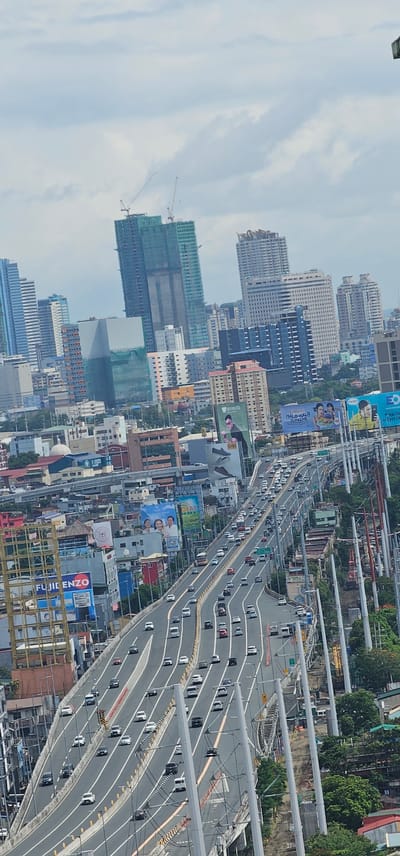 An aerial shot shows a busy elevated highway in Makati, Philippines, choked with traffic under a cloudy sky. The highway, with multiple lanes, is filled with cars and trucks. Surrounding the highway are numerous buildings of varying heights, some under construction, including skyscrapers and mid-rise structures. Billboards are prominently displayed on the sides of buildings and along the highway. Vegetation is visible on the right side of the image, peeking out from behind the buildings and highway infrastructure. The perspective suggests the photo was taken from a high vantage point, possibly another tall building, looking down at the urban landscape.