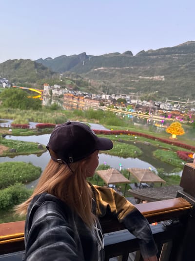 A person wearing a dark baseball cap and a plaid shirt is looking at a scenic view in Zhuoshui, China. The landscape includes a town nestled against a mountain range, a river winding through the town, and decorative structures, including covered walkways and colorful light displays. The person is positioned in front of a window or railing.