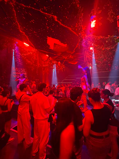The image depicts a dimly lit, vibrant scene at an outdoor event, possibly a music festival or a night market, in Patong, Thailand. The setting is richly decorated with lush green foliage and illuminated by intense red and purple lights, creating a dynamic and atmospheric environment. Several people are present, engaged in various activities, some appearing to be socializing or watching a performance. The overall mood is energetic and festive, suggested by the bright lighting and the density of the crowd.
Notable details include a neon sign that reads "MUSIC SOUNDS BETTER WITH U," which is a prominent element in the scene. The time of day is clearly night, given the artificial lighting and the darkness of the background. The weather appears to be suitable for an outdoor event, with no indications of rain or harsh conditions. The presence of the neon sign and the general ambiance suggest a location that caters to nightlife and entertainment. The people in the image are diverse, with some individuals clearly visible in the foreground and others forming a larger crowd in the background. Their attire and postures suggest a relaxed and celebratory atmosphere. The vibrant red and purple lighting casts a warm, intense glow over the entire scene, highlighting the textures of the foliage and the forms of the people. The composition of the image, with its strong contrasts and bold colors, emphasizes the lively and immersive nature of the event.
