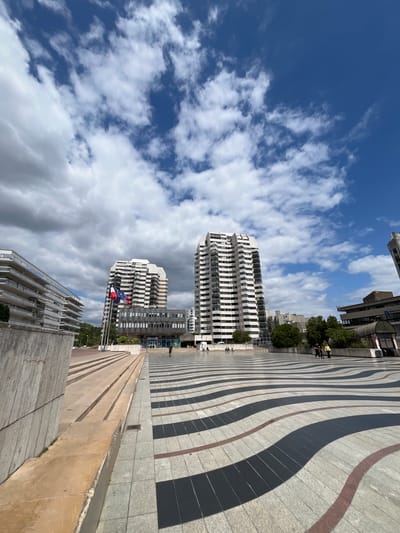 The image captures an outdoor scene with a modern architectural style, likely in Créteil, France. The foreground features a patterned pavement with wavy lines of black, grey, and reddish-brown tiles, leading towards a concrete structure with multiple steps. Beyond this, two tall, white, multi-story buildings with numerous windows stand against a bright blue sky dotted with fluffy white clouds. Flags, including what appears to be the European Union flag and the French flag, are visible near one of the buildings. Trees and other buildings are present in the background, suggesting an urban or semi-urban environment. A few people are seen walking on the pavement and near the buildings. The lighting suggests it is daytime, likely mid-morning or afternoon, with clear, partly cloudy weather. The overall impression is of a clean, contemporary public space. There is no visible text in the image.