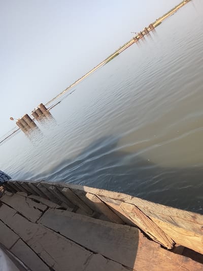 The image displays a daylight scene on a body of water in Ibi, Nigeria, likely the Benue River. The immediate foreground features the exposed interior structure of a wooden vessel, possibly a boat or raft, constructed from unpainted, weathered planks. A small area of blue and white patterned fabric is visible in the bottom left. The water, appearing grey-green with subtle ripples, occupies the midground and reflects the pale sky. In the distance, multiple dark, cylindrical and columnar structures project from the water, consistent with bridge piers or foundations under construction. Lighter materials, potentially scaffolding or formwork, are attached to these structures. A narrow strip of light-brown land defines the distant horizon against a clear, pale blue sky.