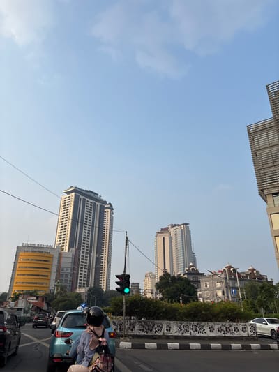 In South Jakarta, Indonesia, on a clear day with a pale blue sky, traffic is at a standstill. A blue car with a rider wearing a light blue top, hijab, and helmet is in the foreground. The rider is facing away from the camera, and their backpack is visible. A traffic light displays a green signal. In the background, the Binus School, a yellow, multi-story building, is visible, as are several tall skyscrapers and other buildings. The environment is urban, with power lines crisscrossing the sky and trees lining the street.