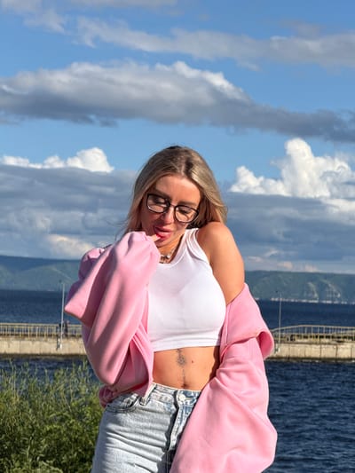 A young woman with blonde hair and glasses is pictured outdoors. She is wearing a white crop top, a pink zip-up hoodie, and light-wash jeans. A tattoo is visible on her stomach. The woman is looking down, with one finger near her mouth, and appears relaxed or contemplative.
The background consists of a bright blue sky with fluffy white clouds, suggesting a sunny day. Beyond the sky and clouds, a body of water is visible, with a distant shoreline or hills. There is also a glimpse of green foliage in the foreground and a concrete structure or bridge stretching along the water's edge.
The orientation of the image is portrait, with the subjects and scene appearing sideways. The lighting indicates it is daytime, likely afternoon given the bright sunlight and blue sky. The overall mood is casual and summery. No text is visible in the image.