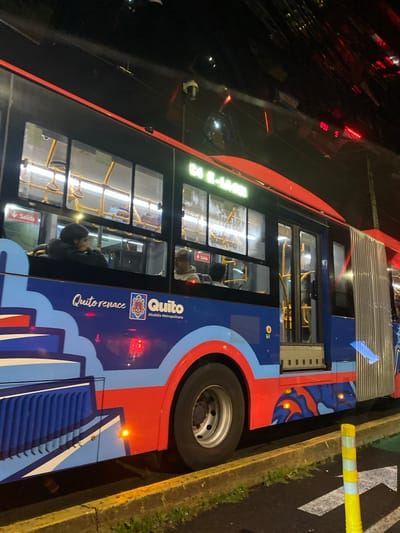 The image depicts a blue, red, and light blue articulated public transport bus stopped at night in Quito, Ecuador. The bus bears the livery "Quito renace" and "Quito Alcaldía Metropolitana" with the city's coat of arms. An electronic display on the front of the bus, partially visible, indicates "E4" and possibly "S-MAGN". Inside the brightly lit interior, several passengers are visible through the windows, some seated, one looking downwards. Yellow grab handles and "Salida" (Exit) signs are present.
The bus exterior features a prominent red stripe above the windows and various light blue and white stylized patterns. Orange side marker lights are illuminated along the lower body. A rear wheel is visible, as is the accordion-style joint of the articulated bus.
The bus is parked alongside a curb painted bright yellow. The ground surface is dark asphalt with a white arrow marking, and a yellow-and-white striped flexible bollard stands in the foreground to the right. The background is dark, indicating nighttime, with various red light reflections and minimal ambient illumination. A security camera is mounted on the upper exterior of the bus.