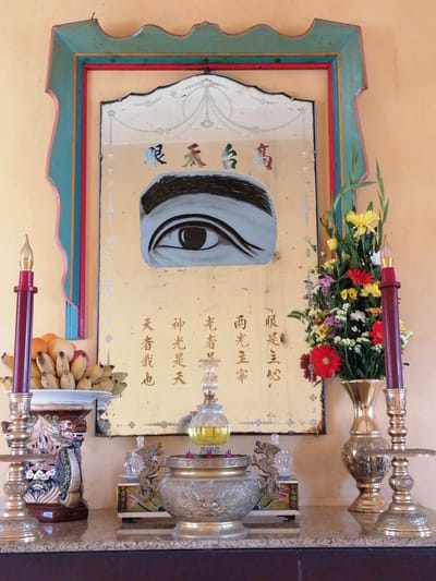 A close-up, head-on shot captures a shrine in Xuân Thành, Vietnam, featuring a collection of religious and decorative items arranged on a granite surface. Dominating the background is a large, ornate mirror with a scalloped, multi-colored frame, painted with an eye motif and possibly Chinese calligraphy. To the left, a red candle in a brass holder stands beside a bowl of fruit, including bananas and oranges, atop a dragon-shaped decorative base. To the right, a brass vase holds a vibrant bouquet of flowers. A second red candle in a brass holder is positioned in front of the flower arrangement. In the foreground, an incense burner, also made of ornate brass, sits between two decorative objects, one appearing to be a small dragon figurine on a box, and the other a clear glass bottle. The wall behind the shrine is painted a pale yellow.