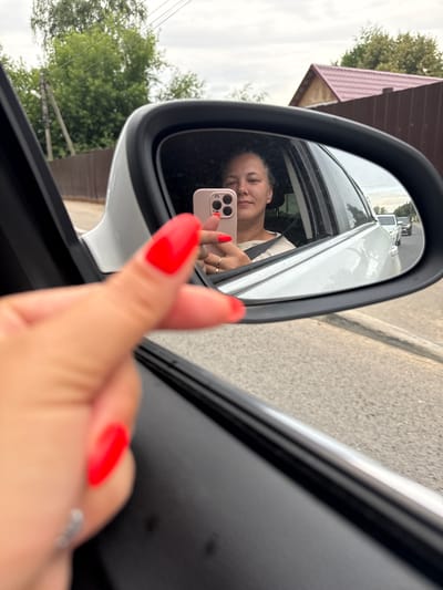 In this media file, a woman is captured taking a selfie in the side mirror of a car. She is sitting in the driver's seat, with her face reflected in the mirror. She is holding a pink smartphone, with the camera facing her. Her nails are painted a vibrant red. In the reflection, she is smiling, and appears to be in good spirits. The background in the mirror shows a street with cars, and some trees and buildings. The exterior of the car is white. The scene appears to be set during the daytime. The location context provided is Dmitrov, Russia. There is no visible text in the image.