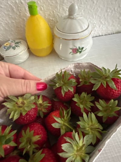 The image displays a close-up, overhead view of several objects arranged on a textured, light-colored surface. The main subjects are a basket overflowing with ripe strawberries and several porcelain decorative items.
The strawberries are vibrant red, with prominent green leafy tops, and appear to be freshly picked and arranged in a woven basket with a textured interior. To the left of the strawberries, there are three porcelain items. The topmost is a lidded jar or pot, white with a gold band and a small floral decal featuring pink roses. Below this is a yellow, elongated object with a green cap, resembling a toy or a dispenser, positioned horizontally. Underneath the yellow object is a small, round porcelain container with a lid, also adorned with a floral decal of roses and other small flowers.
A hand with bright pink nail polish is visible in the lower right quadrant, reaching towards the strawberries, suggesting someone is about to select or handle them. The lighting suggests an indoor setting, possibly natural light, and the overall mood is domestic and summery. There is no visible text in the image. The context provided indicates the location as Krefeld, Germany.