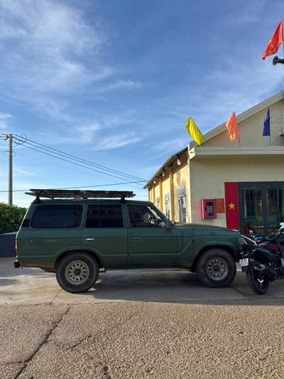 A weathered, olive-green Toyota Land Cruiser is parked on a concrete surface in front of a light yellow building. The vehicle has a roof rack loaded with metal bars. A black motorcycle is parked to the right of the Land Cruiser. The building has a slanted roof with several flags flying, including a red one with a star on the right side, and other colored flags on poles. The sky is blue with scattered white clouds. A utility pole with power lines is visible to the left of the building. The scene appears to be in Trường Thọ, Vietnam, suggested by the text on the building, which includes Vietnamese characters and a license plate on the motorcycle.