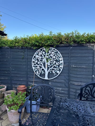 The image captures an outdoor garden or patio scene, featuring a dark wooden fence, lush greenery, decorative elements, and garden furniture under a clear blue sky. The photograph is oriented vertically.

A prominent feature is a tall, black-painted wooden fence, showing some signs of wear and weathering. Along the top of the fence and cascading down the left side is a dense growth of vibrant green foliage, appearing to be a creeper or climbing plant.

Centered on the fence is a large, circular, white metal wall decoration, intricately cut to depict a "tree of life." This decorative tree design includes numerous branches adorned with small, stylized white flowers and light blue-green leaves, standing out against the dark fence.

To the right, black, ornate metal garden furniture is partially visible. The patterned surface of a round or square table occupies the upper right corner, and two matching garden chairs, characterized by their scrolled and lattice designs, are positioned to the right and bottom right.

In the bottom right foreground, several potted plants are arranged. These include a variety of terracotta and ceramic pots of different sizes and finishes, holding leafy green plants, notably a substantial plant with broad green leaves in a large, pale ceramic pot. A grey plastic watering can, partially obscured by the furniture, is visible with some faded red lettering on its side, possibly "HEINZ CO."

The upper left section of the image showcases a bright, cloudless blue sky, crossed by two thin, dark overhead power lines or cables. The bright, even lighting indicates a sunny day, likely morning or afternoon. No people are present in the scene, which conveys a sense of peaceful, well-tended outdoor space.