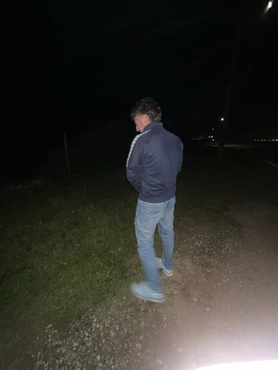 A person in jeans and a blue jacket lies on a patch of ground, which appears to transition from grassy to gravel-like texture. The photo is taken at night or in dim light, with streetlights dimly visible in the background. The person's face is tilted down. A bright light source is at the top right edge, casting a glare.