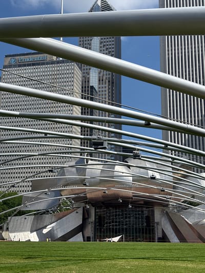 This image shows an outdoor scene in Chicago, USA, with a focus on architectural elements and a clear blue sky. The foreground is dominated by large, curved, silver-colored metal tubes that form an abstract structure. These tubes are arranged in a dynamic, layered pattern, creating a sense of depth and movement.
Behind this metal framework, tall buildings rise. One building, prominently visible, features a repeating pattern of dark windows against a lighter facade. Another building, partially obscured, shows horizontal banding. A closer inspection of one of the buildings reveals the word "Prudential" in large blue lettering, along with a circular logo.
The sky above is a vibrant blue, with a small, white object that appears to be a distant bird or drone. To the right of the image, a section of bright green grass stretches vertically, suggesting a park or open green space adjacent to the buildings and architectural installation. The overall impression is one of modern urban design, with a blend of geometric buildings and flowing metal structures set against a bright, sunny sky. The lighting suggests it is daytime, likely on a clear day, with the sun casting distinct shadows and highlighting the metallic surfaces. There are no people clearly visible in the image, and no specific event is depicted.