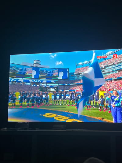 The image shows a screen displaying a soccer match. A group of people, likely a soccer team and staff, are standing on a grassy field. Some individuals are wearing blue and white uniforms, while others are in yellow or blue athletic attire. A large blue and white flag is being held in the foreground, obscuring some of the people. In the background, a stadium is visible with spectators in the stands. A red and white graphic with Cyrillic text and the number "1" is present in the upper right corner of the screen, indicating a live broadcast. The scene is consistent with a sporting event.