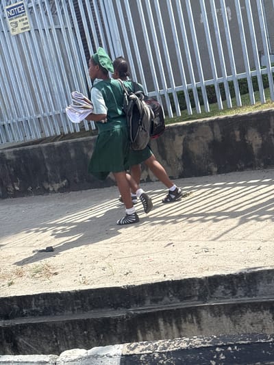 The image depicts two young individuals, likely students, walking on a dirt and concrete pathway in Itirin, Nigeria, on a sunny day. The individual in the foreground wears a green head covering, a light-colored top, a dark green skirt, white socks, and black sandals with white stripes, while carrying a stack of papers or books in their left arm. The second individual, partially obscured behind the first, wears a similar dark green uniform and carries a dark grey backpack with red accents, also wearing white socks and dark sandals. Both are moving from left to right.

To the left and rear of the subjects is a tall, grey metal fence composed of vertical slats, to which a blue "NOTICE VIDEO SURVEILLANCE IN USE ON THESE PREMISES" sign with a camera icon is affixed. Behind the fence, green vegetation is visible. A low, dark grey concrete wall or curb runs parallel to the path, separating it from a lower area or drainage ditch. Elongated shadows from the fence and the individuals are cast on the ground. A small, dark piece of debris lies on the pathway.