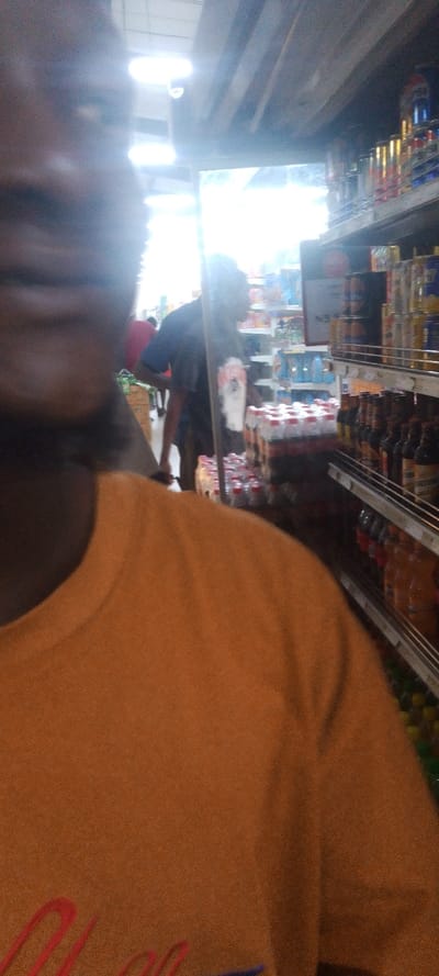 The image shows a person wearing a brown shirt standing in a store aisle in New Karu, Nigeria. The person is looking at a man in a dark blue shirt who is standing in the aisle, in front of a refrigerated display case containing bottles of soda. The store aisle is lined with shelves stocked with various grocery items, including canned goods and beverages. The lighting in the store is fluorescent and the ceiling is concrete. The store is likely to be a supermarket or a grocery store. The image was taken at a location in New Karu, Nigeria, with coordinates (latitude: 8.991, longitude: 7.57).
