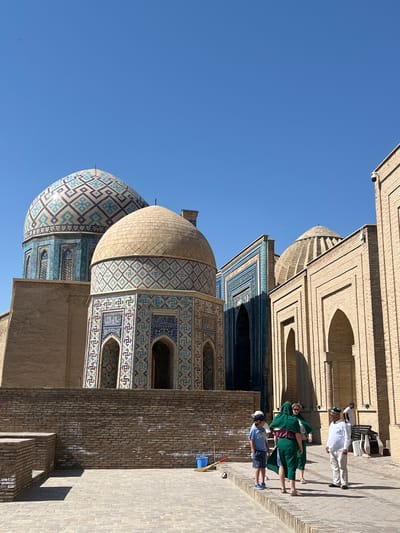 This is a photograph of the Shah-i-Zinda necropolis in Samarkand, Uzbekistan, taken on a sunny day under a clear blue sky. The foreground features a paved courtyard area with a low brick wall. Several people are present: a young boy in a blue t-shirt and shorts, a woman in a green headscarf and dress, and two men, one in a white shirt and light-colored pants, and another in a white shirt and dark pants. They appear to be standing and conversing. In the midground, the iconic blue and turquoise tiled domes and facades of mausoleums are visible, characterized by intricate geometric patterns and arched doorways. The architecture is a striking example of Islamic art and architecture. The background is dominated by the vast expanse of the blue sky, with a single small bird visible in the upper left quadrant. The overall impression is one of historical grandeur and cultural significance.