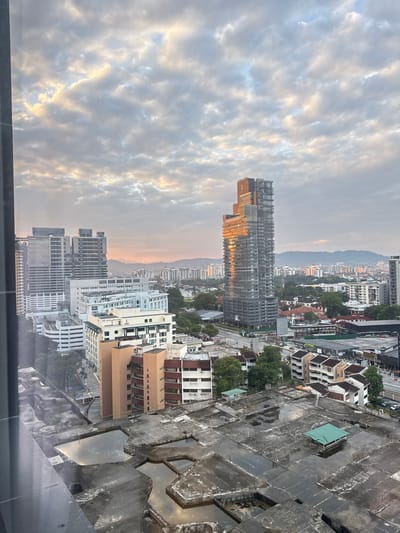 The image captures a panoramic view of Kuala Lumpur, Malaysia, likely taken during sunrise or sunset. In the foreground, a weathered, unfinished concrete structure with several large, water-filled cavities dominates the lower half of the frame. Numerous rebar protrusions suggest ongoing or halted construction.
Moving upwards, the mid-ground showcases a dense urban landscape with a variety of buildings. Prominent among them is a sleek, modern skyscraper with a distinctive stepped design, its glass facade reflecting the warm hues of the sky, indicating it's catching the golden hour light. Adjacent to this are other contemporary office buildings with extensive glass exteriors and some older, multi-story residential or commercial structures. The architecture suggests a mix of modern development and older urban planning.
In the background, a range of hills or mountains form a hazy silhouette against the sky, softened by distance and atmospheric conditions. The sky itself is filled with a dramatic expanse of clouds, ranging from soft grays and blues to areas illuminated by sunlight, creating a textured and dynamic ceiling to the cityscape. The overall impression is that of a bustling metropolis viewed from a high vantage point, with the natural elements of the sky and distant landscape providing a contrasting backdrop to the built environment. There are no visible individuals or discernible human activity in the frame, focusing solely on the architectural and atmospheric elements.