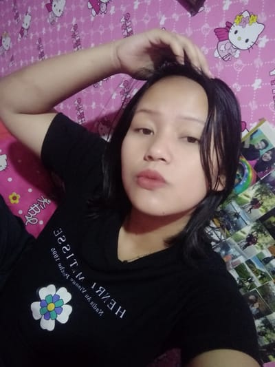 A young woman is captured in a selfie. She wears a black t-shirt featuring the text "HENRI MATISSE" and a flower graphic. Her dark hair is shoulder-length. She is positioned against a background of pink Hello Kitty patterned bedding and a collage of photographs. One hand is raised and touches her hair. She has a neutral expression and is looking directly at the camera.