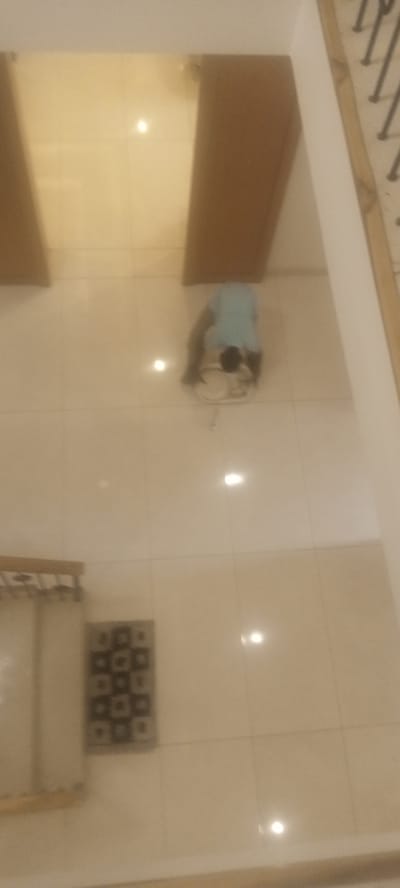 A person is kneeling and cleaning the floor in what appears to be an indoor tiled space in Unguwar Rimi, Nigeria. The space contains two brown wooden doors on the upper portion of the image. A dark patterned rug and a partially visible piece of furniture are located at the bottom left of the image. A section of staircase railing is visible on the right.