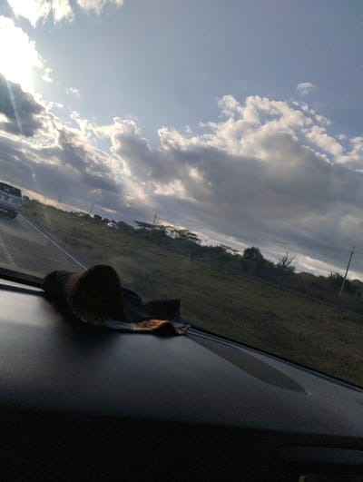 The image is taken from the interior of a car, looking out the windshield toward the road ahead. The car is stopped or slowly driving, and a rag sits on the dashboard. The view shows a partly cloudy sky, with the sun shining behind the clouds. There is a two-lane paved road with a vehicle in the distance ahead. The road is lined with trees and brush. The image is taken at coordinates -1.495, 37.065, which places the location in the town of Athi River, Kenya.