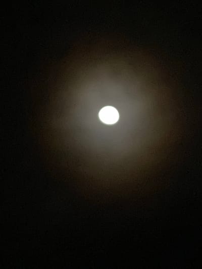 The image shows a bright, full moon centered in the dark night sky. There is a hazy or diffused ring of light around the moon, possibly due to atmospheric conditions. The surrounding sky is dark, with no other visible stars or objects. No specific information about the location in Maigizo, Nigeria can be determined from the image. There are no people, objects, or actions depicted other than the presence of the moon in the night sky.