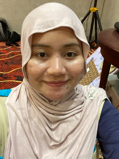 A selfie shows a woman wearing a light pink hijab and a matching scarf. She is smiling and looking at the camera. Her face has some visible blemishes. Behind her, a patterned rug is visible on the floor, along with a black backpack and a tripod. A dark wooden table is to her right, with some papers and other objects on it. The overall lighting suggests an indoor setting. The specific location in Banjarbaru, Indonesia, cannot be determined from the image alone.