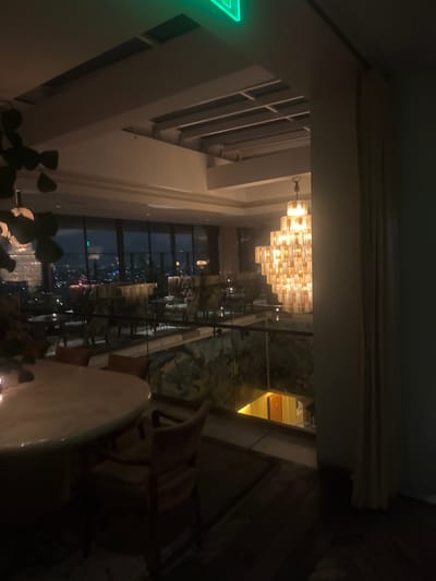 The image shows an interior view of a restaurant or bar located in West Hollywood, California, United States of America, likely at night. A large, multi-tiered chandelier dominates the upper-center of the frame. A vertical panoramic window provides a view of the city at night. Tables and chairs are visible closer to the bottom of the frame, indicating a dining or seating area. A partially visible green exit sign is present on the far left. The overall lighting is dim.