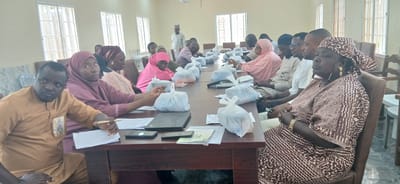 The image shows a group of people, likely attending a meeting or training session, seated around a long table in a room. The subjects are diverse in age and gender, with many wearing traditional African attire, including headscarves and long robes. Several individuals are looking towards the camera or engaging with the person taking the photograph.
On the table, there are several white plastic bags, possibly containing food or materials, along with laptops, mobile phones, papers, and pens. This suggests a formal gathering where participants may be receiving or distributing items, or engaging in work-related activities.
The setting appears to be an indoor meeting room. The walls are painted a light color, and large windows with metal bars are visible, suggesting a potentially secure or utilitarian building. The overall lighting is bright, indicating it is daytime. The presence of a whiteboard or projection screen is suggested by a faint outline on the wall in the background, and a person standing in the background wearing a traditional cap suggests this is likely in Nigeria.
The mood seems to be one of attentiveness and engagement, with some individuals appearing serious and focused, while others have a more relaxed or curious expression. The presence of multiple people in a meeting setting indicates a social or professional event. No specific text is clearly legible in the image.