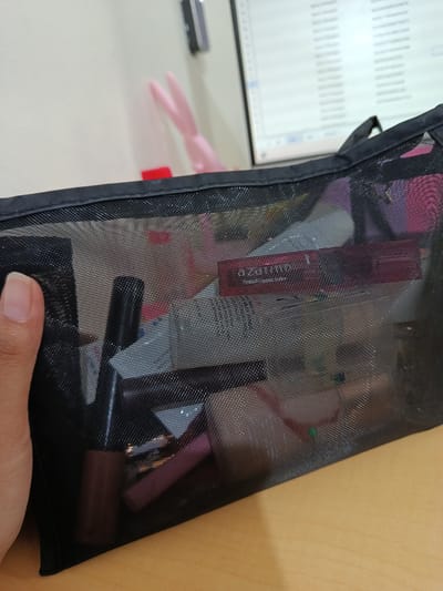 A close-up shot shows a partially filled black mesh makeup bag on a wooden surface. A hand holds the edge of the bag. Visible contents inside the bag include makeup items like lipsticks, mascara, and other cosmetic products. Behind the bag, a computer monitor displays spreadsheet-like data. In the background, there's a pink bunny-shaped object and a red item, possibly a container. The setting appears to be indoors, likely an office or home desk. The city of Bandung City, Indonesia, is only mentioned as a requirement, but there is no evidence in the image that corroborates its location.