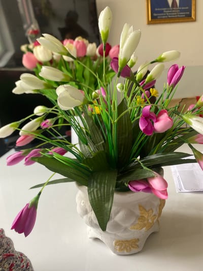 A close-up, eye-level shot features a white ceramic vase containing a bouquet of artificial flowers. The vase is intricately designed with raised, golden floral patterns and sits on a white, reflective surface, likely a table. The bouquet is comprised of various artificial flowers, predominantly white and pink tulip-shaped buds, interspersed with green foliage and a few small yellow accents. The background is blurred but suggests an indoor setting, with a dark rectangular object, possibly a monitor or television, on the left, and a framed picture on the wall to the right. The overall lighting is bright, casting subtle shadows. The scene is devoid of any human presence. Based on the framed picture, which appears to contain official symbols or portraits, and the general ambiance, the location is likely an office or a formal room within the city of Dushanbe, Tajikistan.