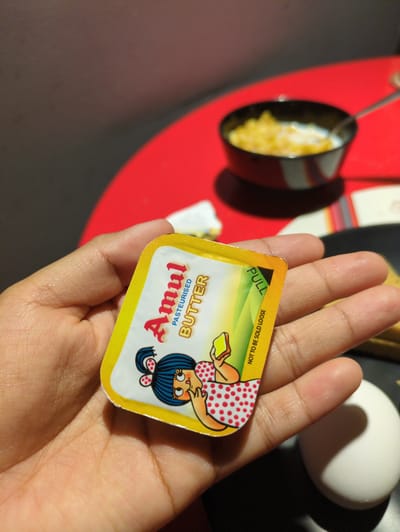 A human hand holds a small, yellow-packaged Amul Pasteurized Butter container. The butter container is in focus and displays an illustration of a cartoon girl in a red polka dot dress. In the background, there is a blurry image of a small pan with cooked food, a white egg, and a package. The image was taken in Bengaluru, India.