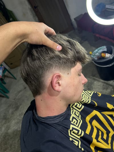 A young male with light brown, textured hair is seated in a dimly lit indoor environment, identified as Hvostiane, Bulgaria, receiving a haircut. His eyes are closed, and he wears a dark blue haircutting cape with a gold geometric pattern. An unseen individual's hand, exhibiting light skin tone, holds a section of the male's hair on the top of his head, pulling it upwards. The male's hair features a faded cut on the sides and back. In the background, a light-colored wall with a partial view of a wooden door or frame is visible. A circular LED ring light, emitting white light, illuminates the scene from the upper right. A black bucket containing a plastic bottle and other indistinct items is present in the right background. Green chairs are partially visible in the far left background, and the floor appears to be a grey, hard surface.