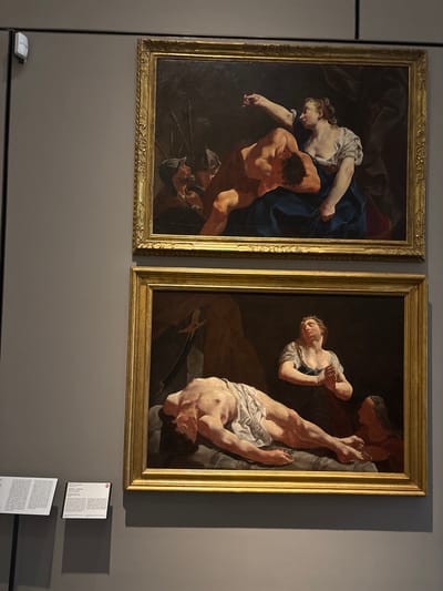 The image displays two large, rectangular oil paintings, framed in ornate gold, hanging side-by-side on a dark grey wall in what appears to be a museum or gallery setting. The ambient lighting is subdued, with spotlights illuminating the artworks and casting soft shadows on the wall.
The painting on the left depicts a dramatic scene with several figures. In the foreground, a muscular man, his back mostly towards the viewer, struggles or interacts with a woman positioned above him. The woman wears a white blouse and dark blue drapery, her arm outstretched. Her expression suggests intense focus or effort. Below these two, faint details of weapons, possibly spears or swords, and armored elements are visible in the dark background, hinting at conflict or violence.
The painting on the right also features multiple figures in a dynamic and dark composition. A man, largely nude except for a white cloth around his lower body, is depicted upside down, his legs extended towards the top of the frame. A woman in a light-colored dress and headscarf supports or holds the man's arm, her face conveying sorrow or concern. Another figure is partially visible in the darker background.
Both paintings exhibit a Baroque style, characterized by intense chiaroscuro, dramatic poses, and rich, somber color palettes. The subjects appear to be entangled in moments of struggle, suffering, or intense emotion.
Below the right painting, two white informational plaques are affixed to the wall. The top plaque clearly displays the artist's name: "GIULIA LAMA". Below the name, titles are visible, including "Giuditta e Oloferne" (Judith and Holofernes), followed by "Maddalena" (Mary Magdalene) and "e altre opere" (and other works). This indicates the paintings are likely by the Venetian Baroque artist Giulia Lama, with at least one possibly depicting the biblical story of Judith and Holofernes. A small white sensor or device is visible on the wall below the left painting.
