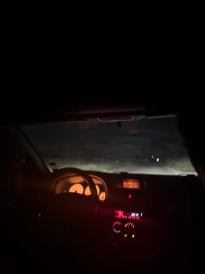 A nighttime view from inside a car. The illuminated dashboard and speedometer are visible. Through the windshield, the road ahead is dimly lit. It's not possible to determine the exact location in Elhovo, Bulgaria without more information. No people are clearly visible.
